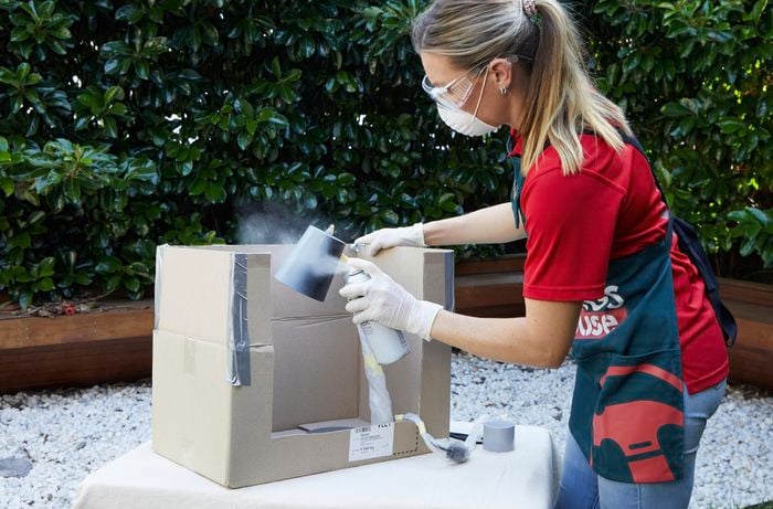 A woman wearing an apron and safety gear spray paints a lamp outdoors.