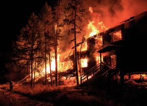 A building and nearby trees engulfed in a large bushfire fire at night, with a firefighter spraying water