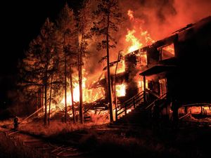 A building and nearby trees engulfed in a large bushfire fire at night, with a firefighter spraying water