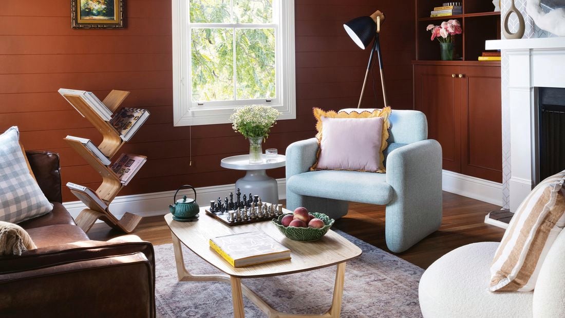 Fruit basket, book and a kettle on a coffee table, blue armchair in the foreground and with yellow flowers in vase on top of a side table next to it.