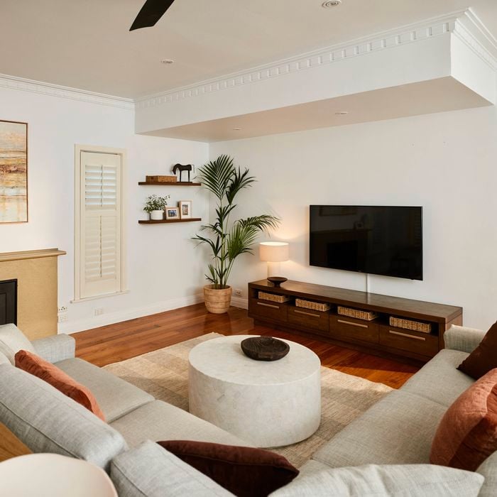 Light-filled living room with wooden floors, a wall-mounted TV above a low timber cabinet with baskets, a round coffee table on a rug, neutral sofas, indoor plants, and minimalist wall shelves.