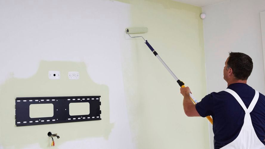 Person in overalls painting a white wall light green with a roller near a TV mount and electrical outlets.