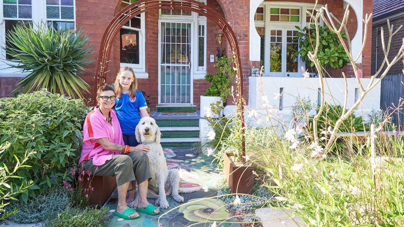 A woman sits on a bench in a front yard with a child and a large white dog beside her. Behind them are some plants, and a red‑brick house with green tiled steps.