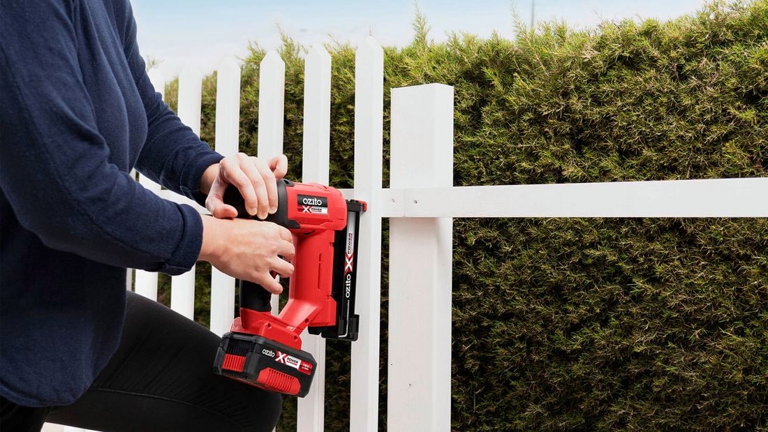 A man wearing a protective safety mask uses a paint spray gun to spray light blue paint onto a white fence.
