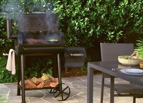  A black offset smoker barbecue with beef brisket and vegetables cooking inside, with firewood stored underneath.