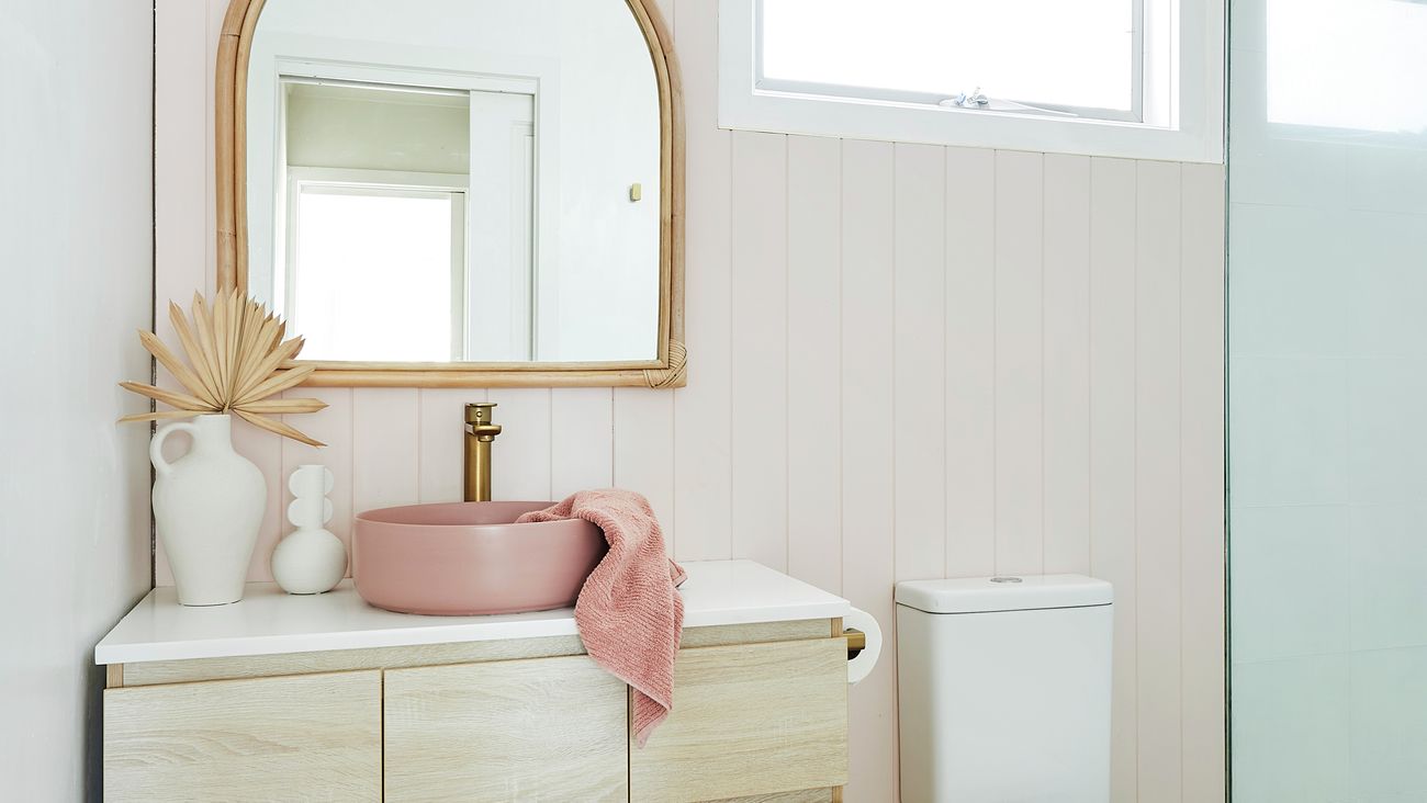 A coastal-themed bathroom vanity with a pink basin, pink towel, and a bamboo-framed arched mirror. A toilet and shower are next to it.