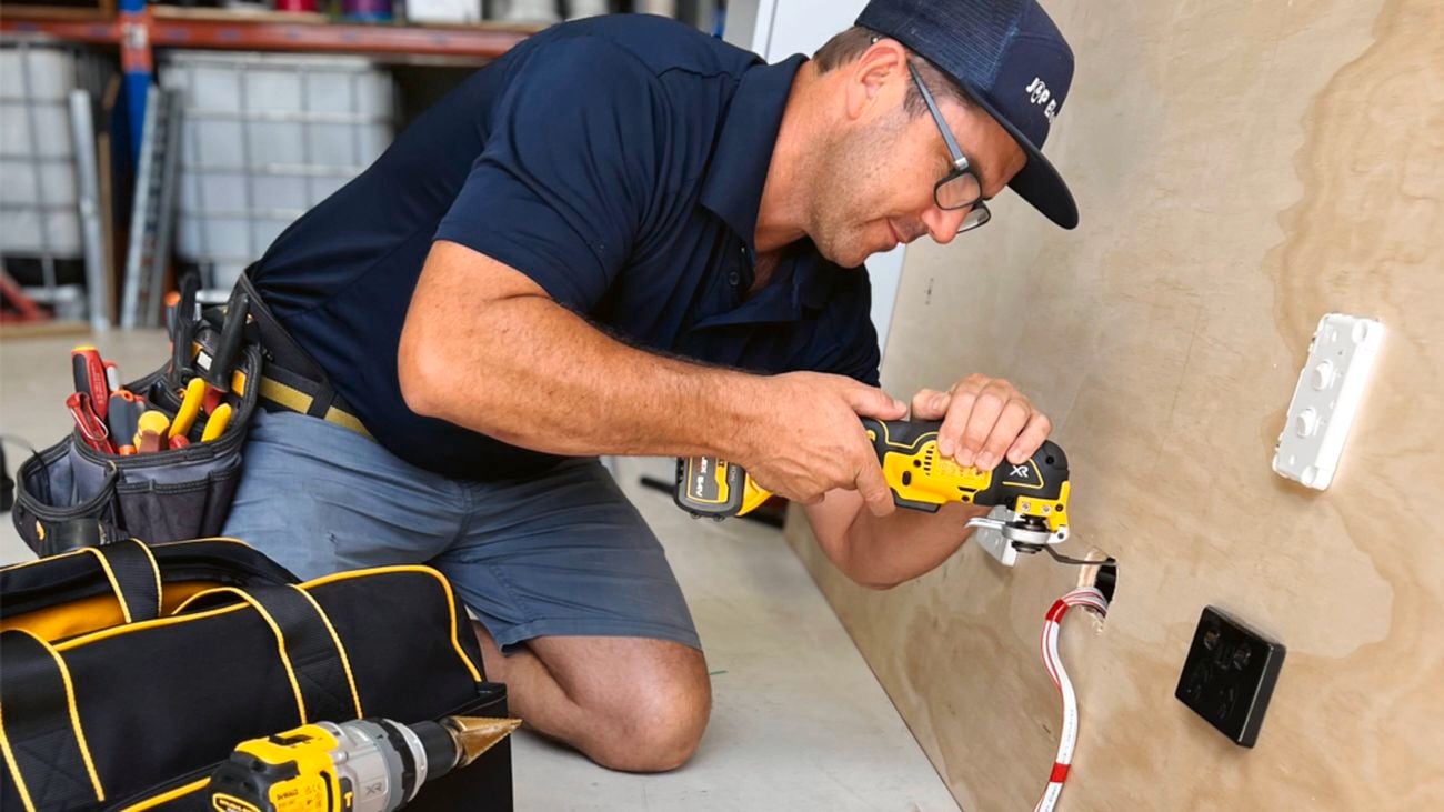 An image of an electrician wearing a blue polo shirt and shorts, glasses and a navy baseball cap uses a DeWALT multitool to carry out electrical work on a wall socket