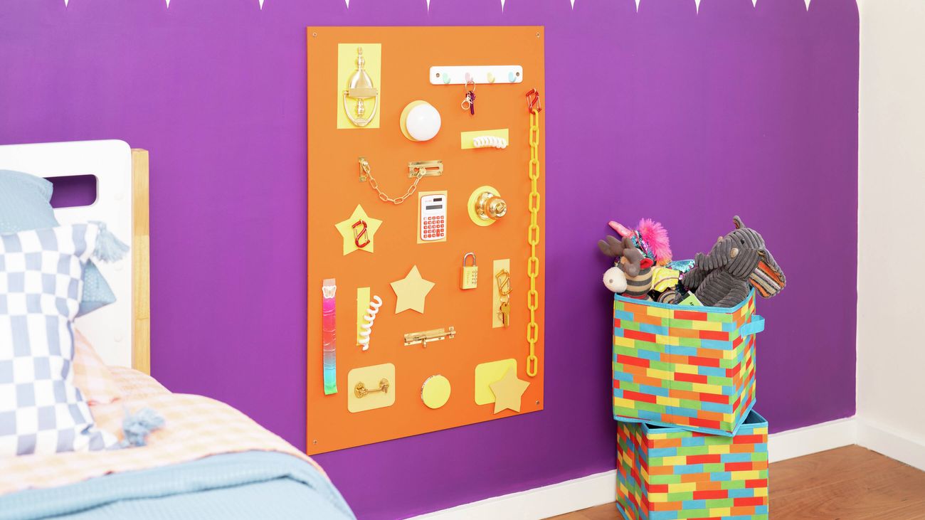 A colourful children’s room with a purple scalloped wall and an orange busy board mounted on it, covered with various tactile objects, beside a box of toys.