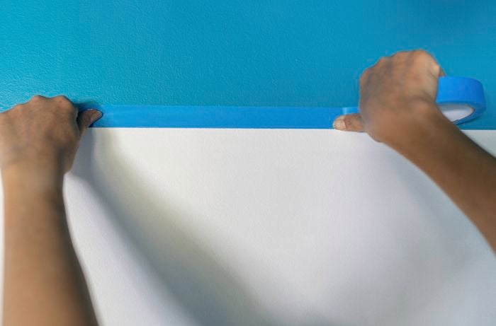 A person carefully applies painter’s tape along the edge of a freshly painted blue colour cap to create a clean, sharp line between the coloured section and the white wall. 