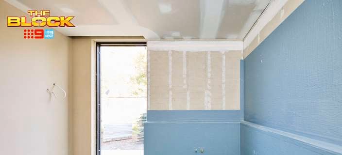 An unfinished bathroom with blue wall panelling, a partially plastered wall, green flooring, and a large floor-to-ceiling window.