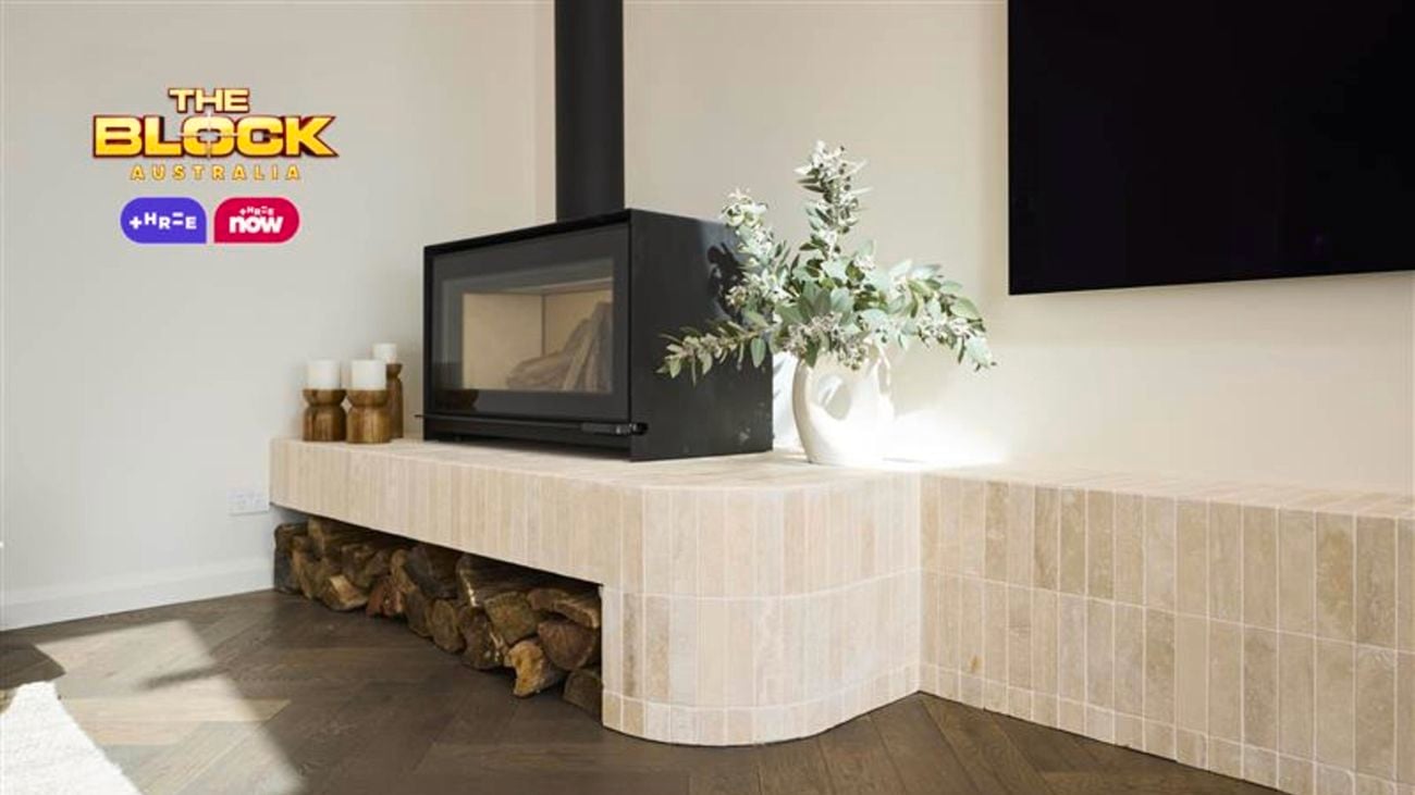 A modern living room corner featuring a black-framed fireplace built into a light-colored tiled structure that extends into a bench-like surface. Firewood is stored beneath the bench, and the top is decorated with a white vase of green foliage and candles. The floor has dark wood herringbone patterning, and a large black screen is partially visible on the wall. 