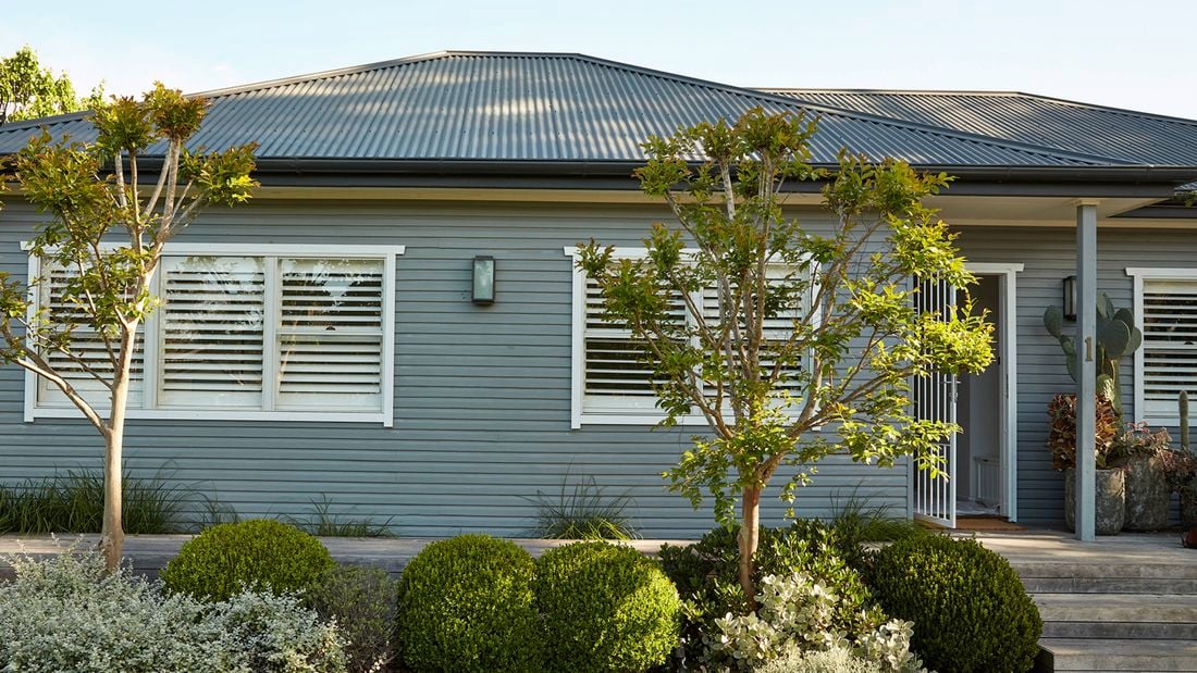 Exterior of a grey coloured weatherboard home with many plants and trees in front of it.