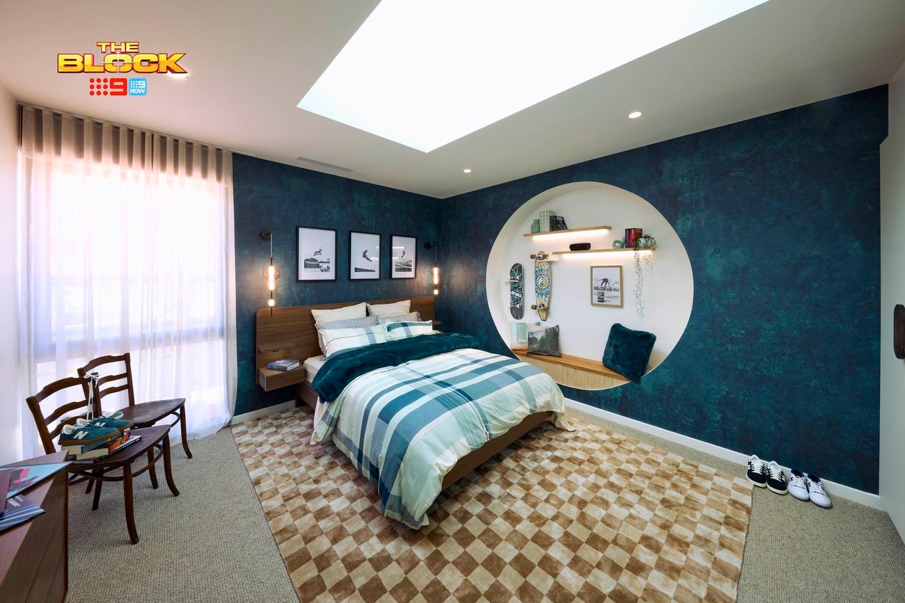 Blue bedroom with a double bed, textured blue wallpaper and blue checked linen. The bed is on a brown checked rug and there is a circular recess in the wall with a bench seat and clothing hooks.