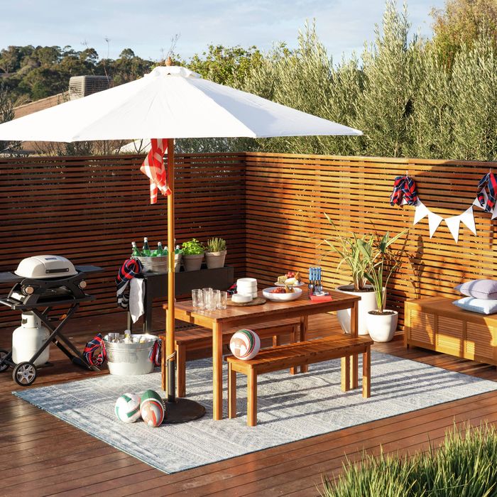 Outdoor entertaining area on a timber deck featuring a large white market umbrella, wooden dining table with bench seating, portable gas barbecue, drinks in ice buckets and decorative plants. The space is styled for backyard entertaining with sporting balls, outdoor cushions and a timber privacy screen surrounded by trees.