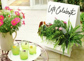 A white planter box filled with green plants and pink flowers, with a white welcome sign behind it that reads "Let's Celebrate". [[2609] MAK...me sign_V1 | Word]