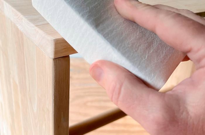 Close up of a hand holding a sanding block with abrasive paper wrapped around it, in front of a timber table