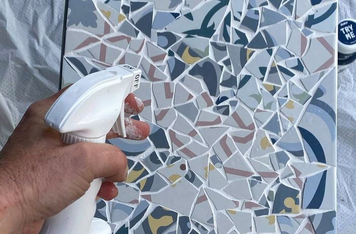 A hand spraying grout sealer onto patterned mosaic tiles.