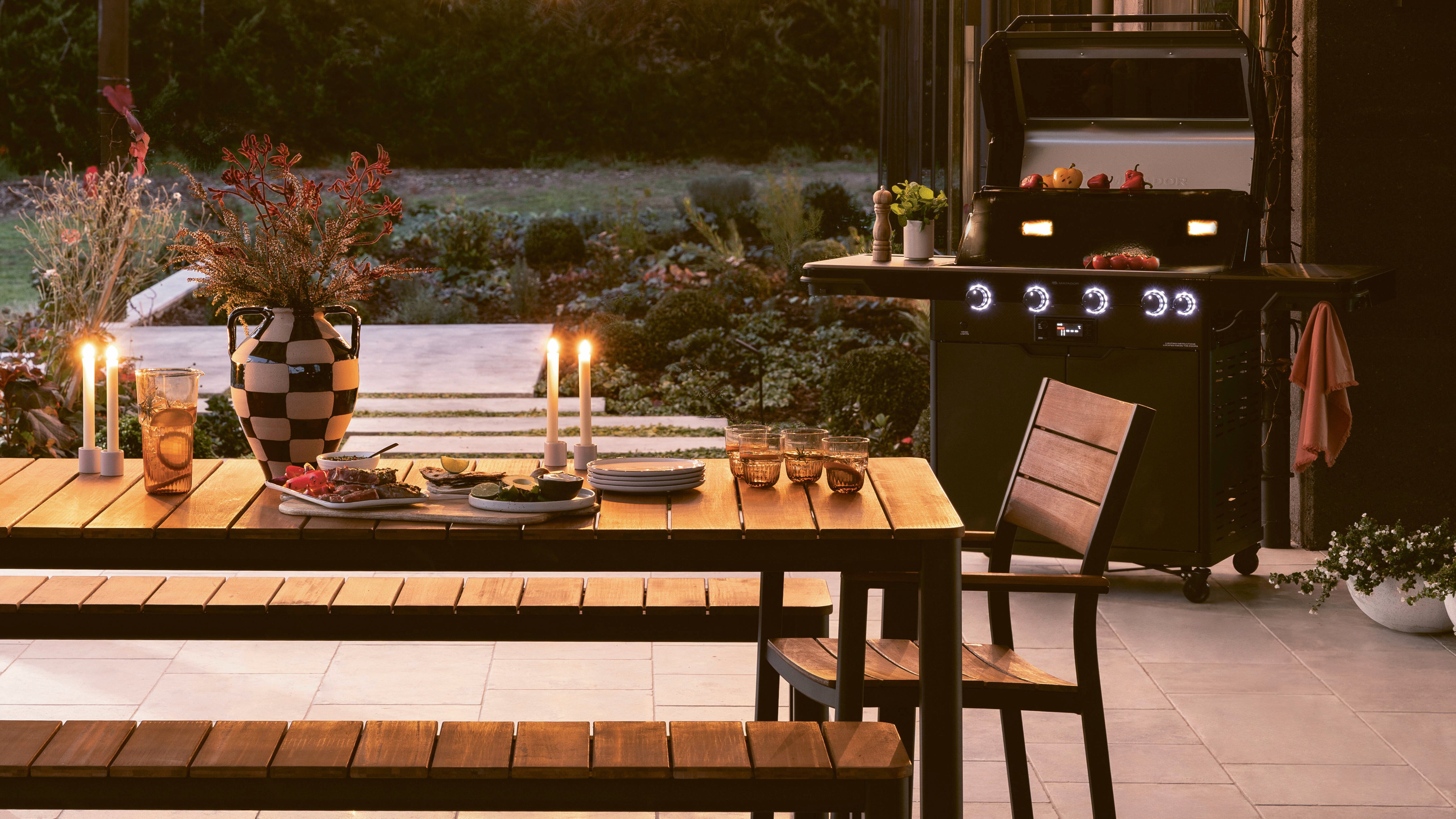 Outdoor patio with a wooden dining table, benches, a chair, string lights, and a barbecue grill.