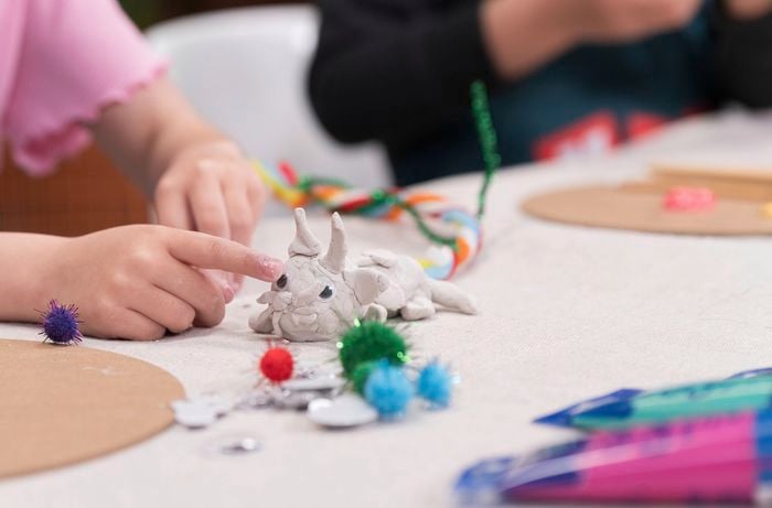 A finger points to a clay creature decorated with googly eyes, surrounded by colorful craft items on a table.