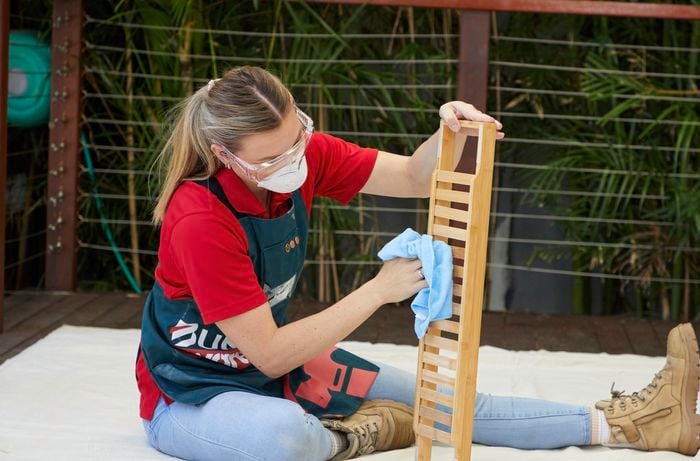 A woman in a Bunnings Warehouse apron sits on a drop sheet and wipes down a bath caddy.