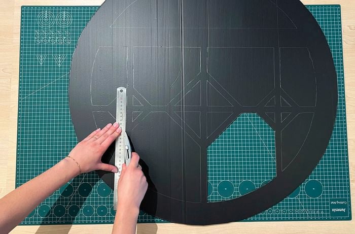A pair of hands use a ruler and utility knife to cut out the marked window on black flute board on a green cutting mat