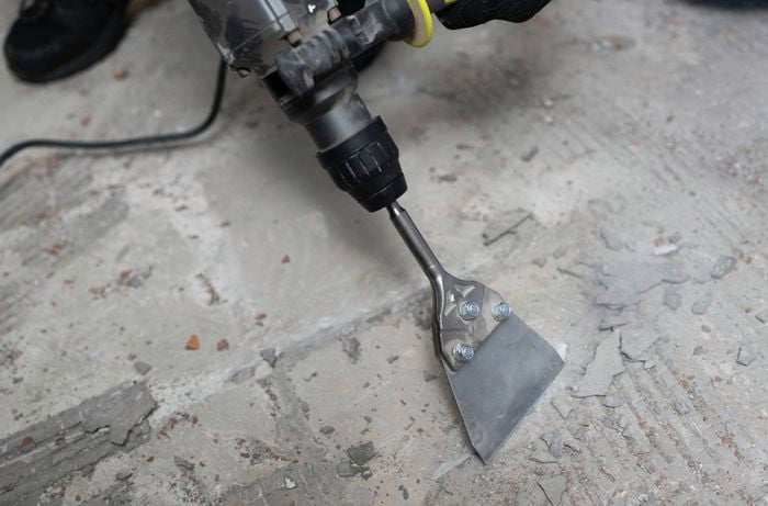 A rotary hammer with a wide chisel attachment is being used to remove tile adhesive and debris from a floor.
