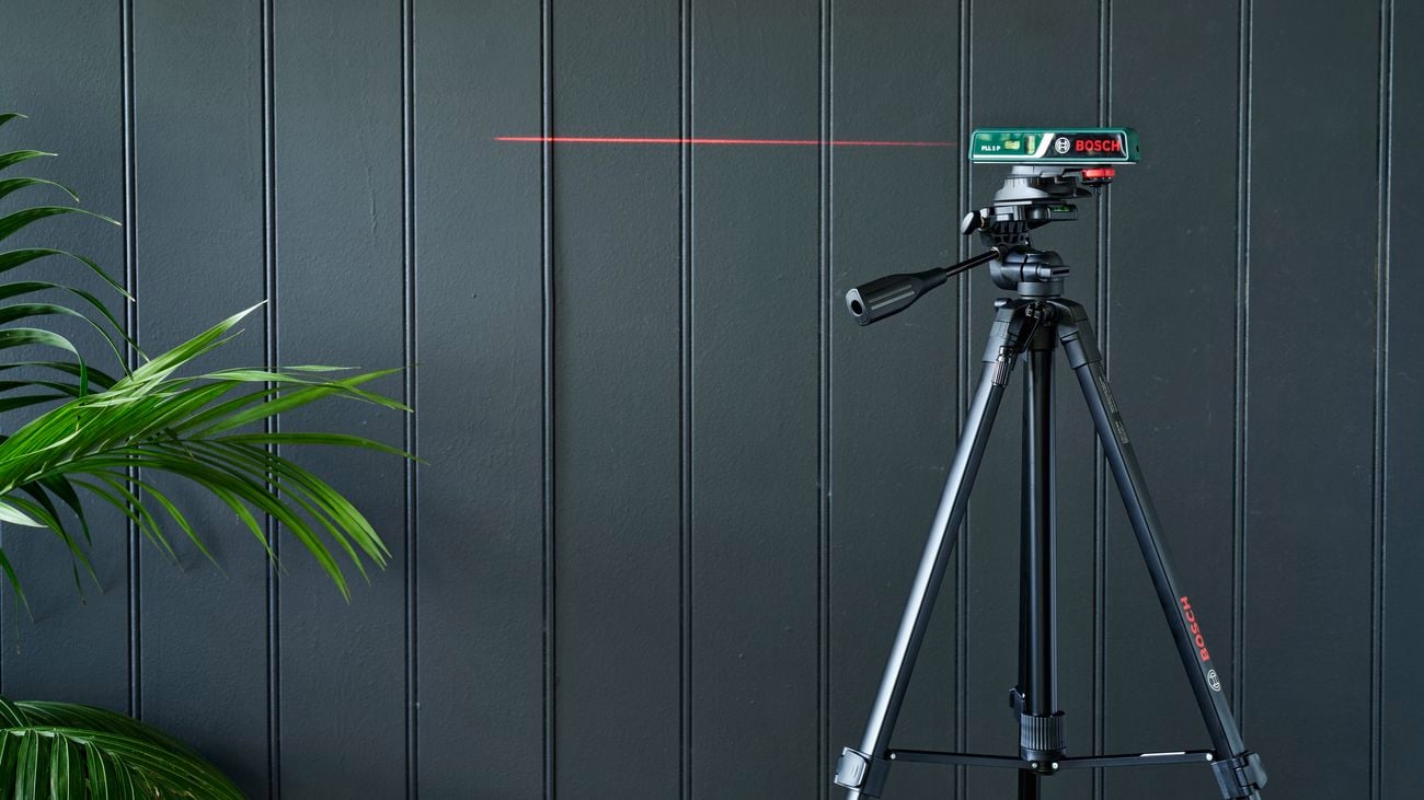 Bosch laser level on tripod pointing onto plain black wall with plant on the left.