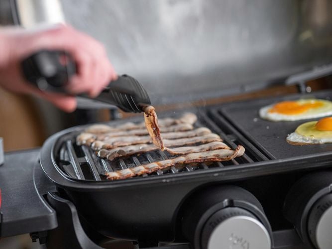 A person cooking bacon and eggs on a BBQ using tongs.