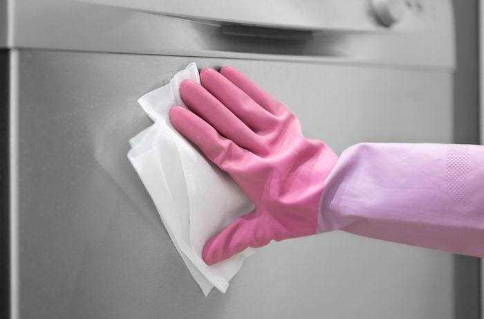 A hand in pink gloves is using a paper towel to clean the stainless steel surface of the dishwasher. 
