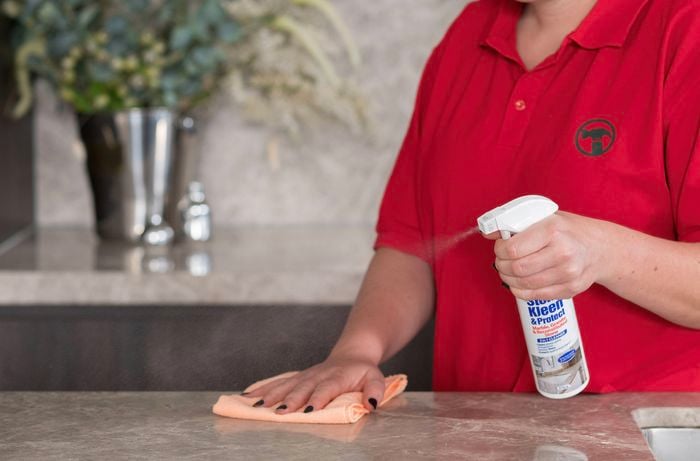 A person in a red T-shirt is using a microfiber cloth to wipe the benchtop with Stone Kleen & Protect Marble Granite Cleaner.