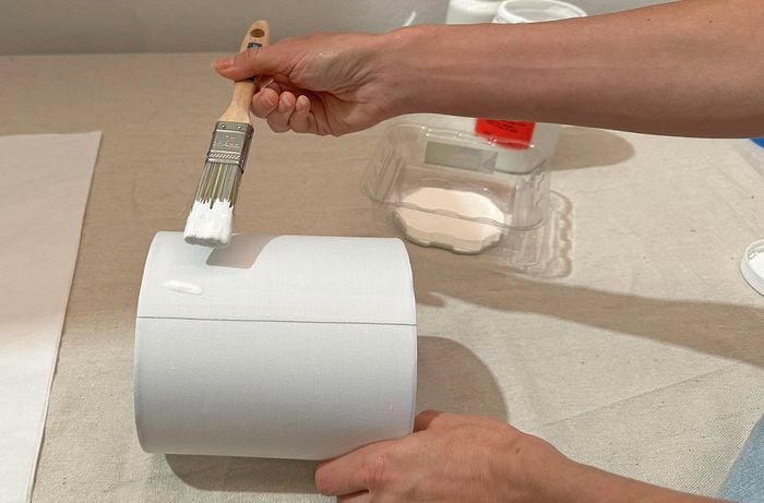 A close-up of a right hand holding a paintbrush and painting a lampshade while the left hand steadies it