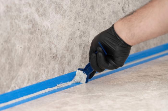 Black-gloved hand uses a blue silicone finishing tool to smooth a bead of silicone along the joint between a stone-look benchtop and matching splashback, with blue masking tape protecting the edges.