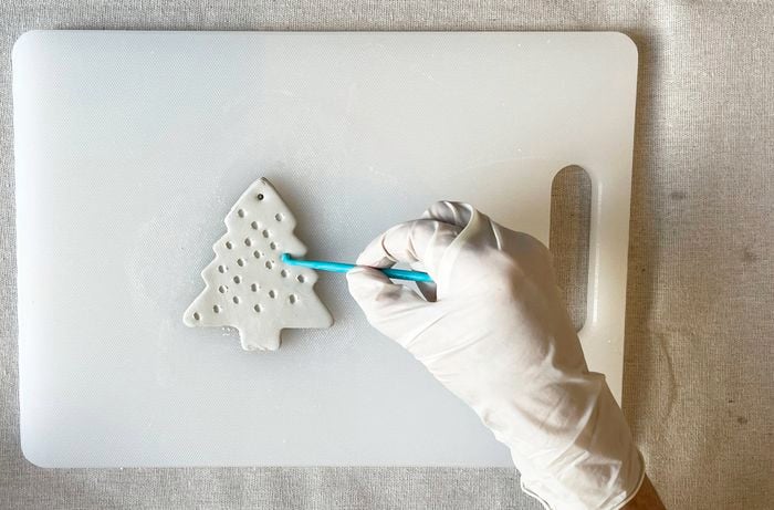  A white gloved hand using a clay tool to make holes in a white clay Christmas tree that sits on a white chopping board.
