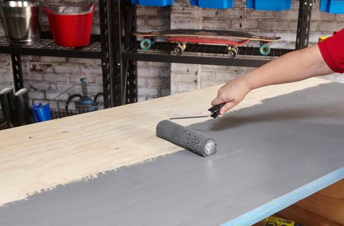 A person painting a plywood benchtop grey using a roller
