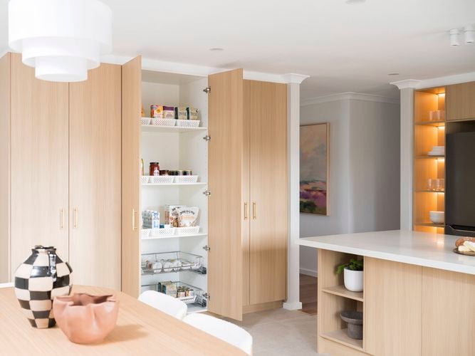 Light brown modern kitchen with a white benchtop, with the pantry double doors open and a table and chairs in it.