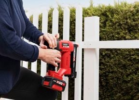 A man wearing a protective safety mask uses a paint spray gun to spray light blue paint onto a white fence.