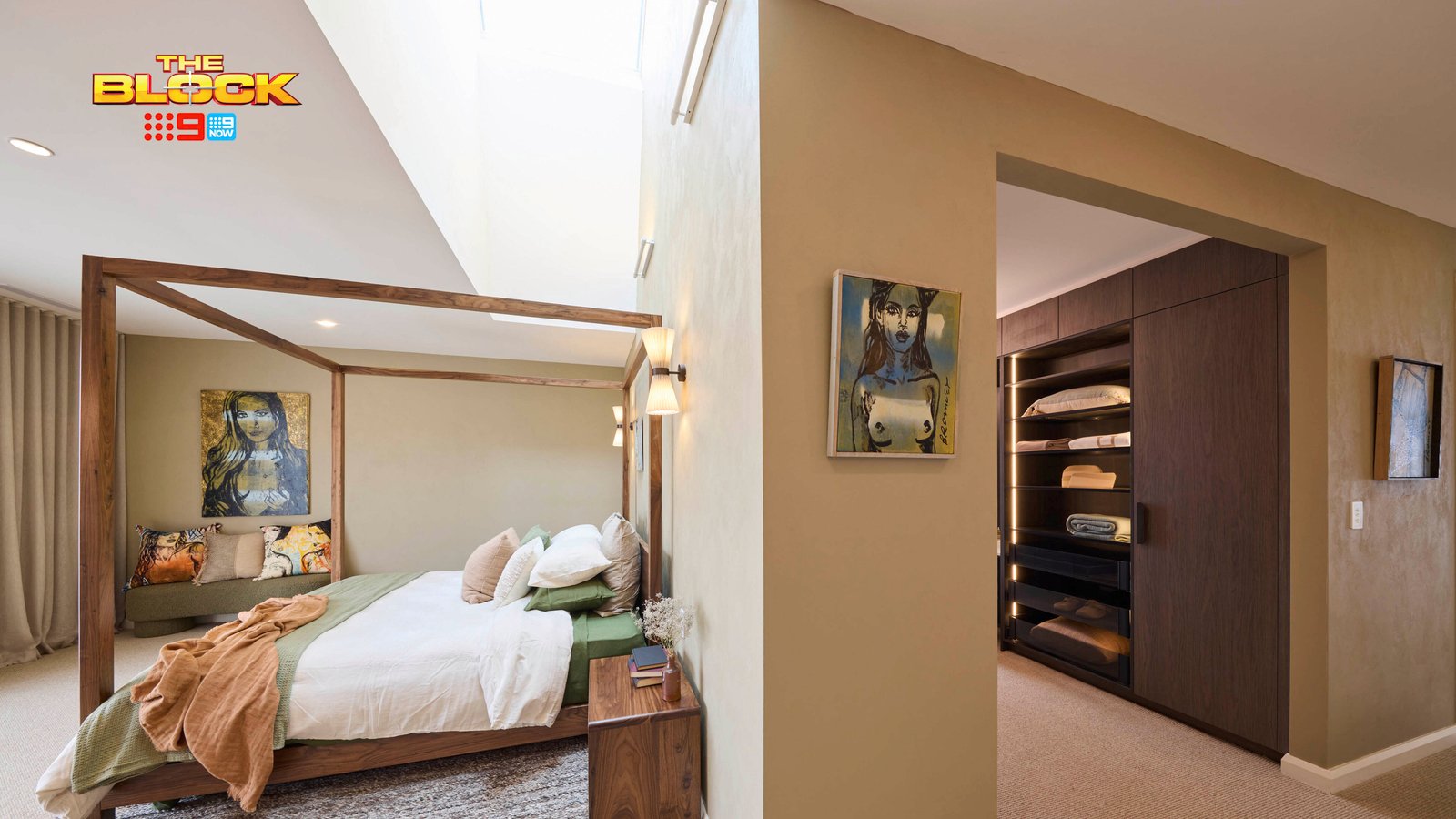 A modern bedroom featuring a wooden canopy bed with white and green bedding. A skylight above the bed allows natural light to fill the room, which has beige walls and carpeted flooring. To the right, an open doorway leads to a walk-in closet with shelves of folded clothes. 