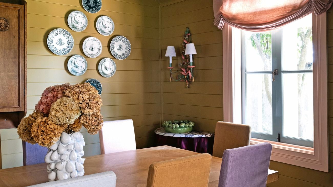 Old style dining room with pale yellow, wooden walls, open orange curtain letting sunlight onto a wooden dining table and chairs.