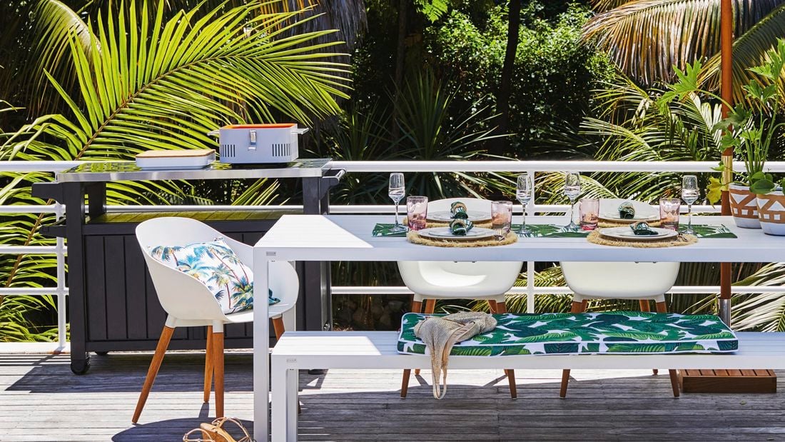 White outdoor table setting with glasses, cushions and pot plants.