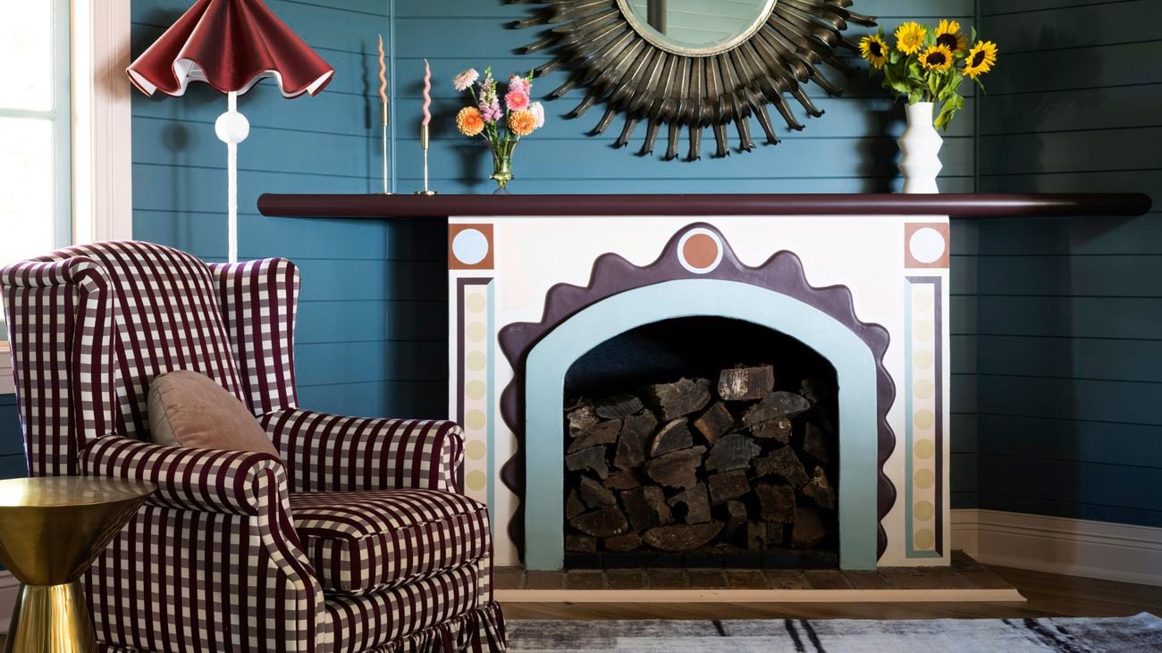 Shot of fireplace with checkered arm chair, blue wall behind and decorative flowers on mantel piece.