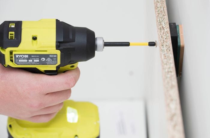 A person is using a power drill to attach the back panel into the wall studs and fix the cabinet in place using screws.