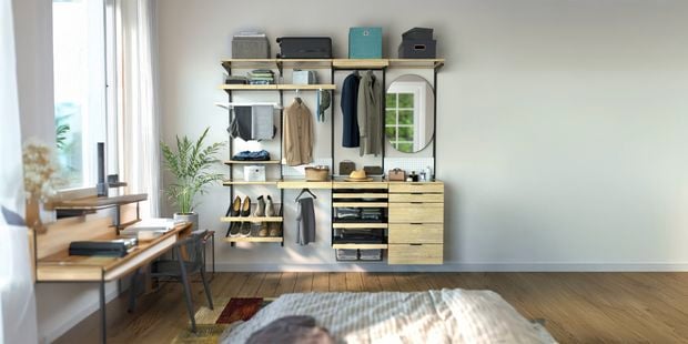 Modern bedroom with a wall-mounted modular wardrobe system featuring wood shelves, hanging rails, drawers, shoe storage, and a round mirror. The organized storage display holds folded clothing, shoes, boxes, and accessories, while a desk sits beside a large window in the foreground.