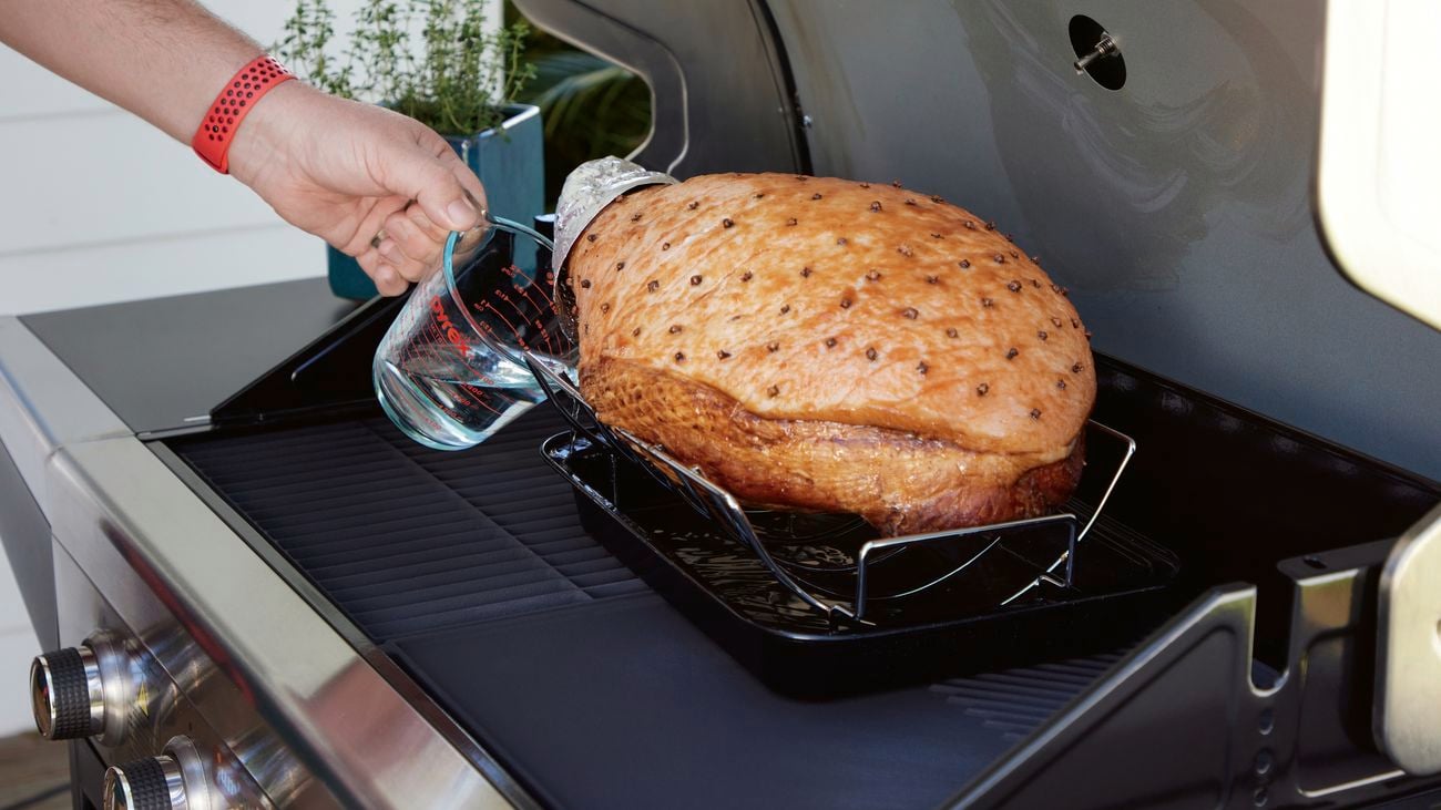 A ham sits in a roasting pan on top of a barbecue with the hood up.
