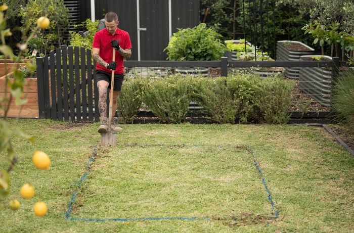 A Bunnings Team Member is using a shovel to dig along the marked outline of a garden bed to prepare the area by clearing away any grass or weeds. 