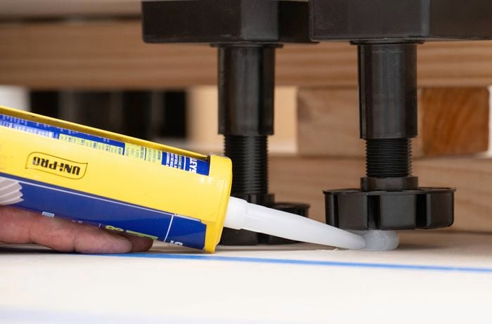A person is applying a silicone sealant with a caulking gun to secure the base of a cabinet feet to the floor.