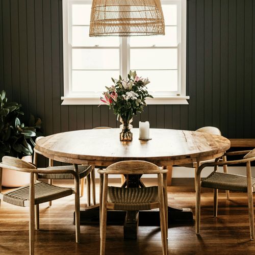 Warm dining room with a round timber table, woven dining chairs, floral centrepiece and large rattan pendant light against a dark feature wall.