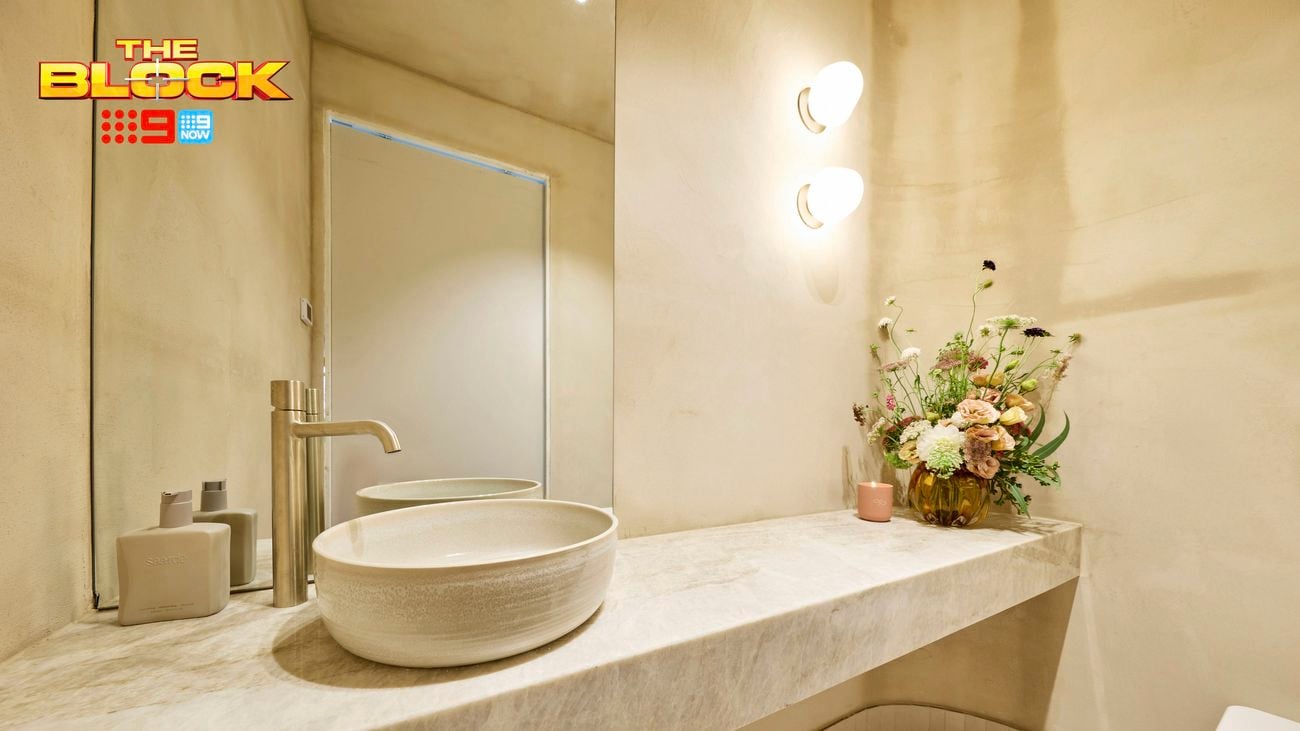 A modern bathroom with a sink on a benchtop with a vase with flowers in it on the benchtop, a mirror and lights on the wall.