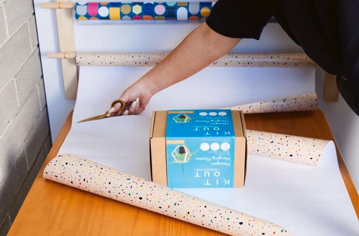 Wrapping paper rolled face-down on a wooden table with a box in the middle. A person is cutting the wrapping paper with scissors.