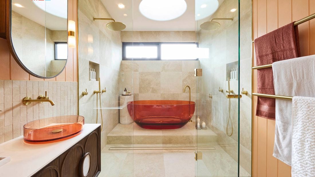 Modern bathroom with amber tub in glass shower, beige tiles, dual overhead showerheads, amber basin with round mirror, and gold towel rack. 