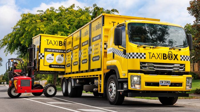 Yellow TAXIBOX truck parked on a suburban street with multiple storage boxes loaded on the back, while a small red forklift lifts another yellow TAXIBOX unit beside the truck. Trees and houses are visible in the background under a partly cloudy sky.